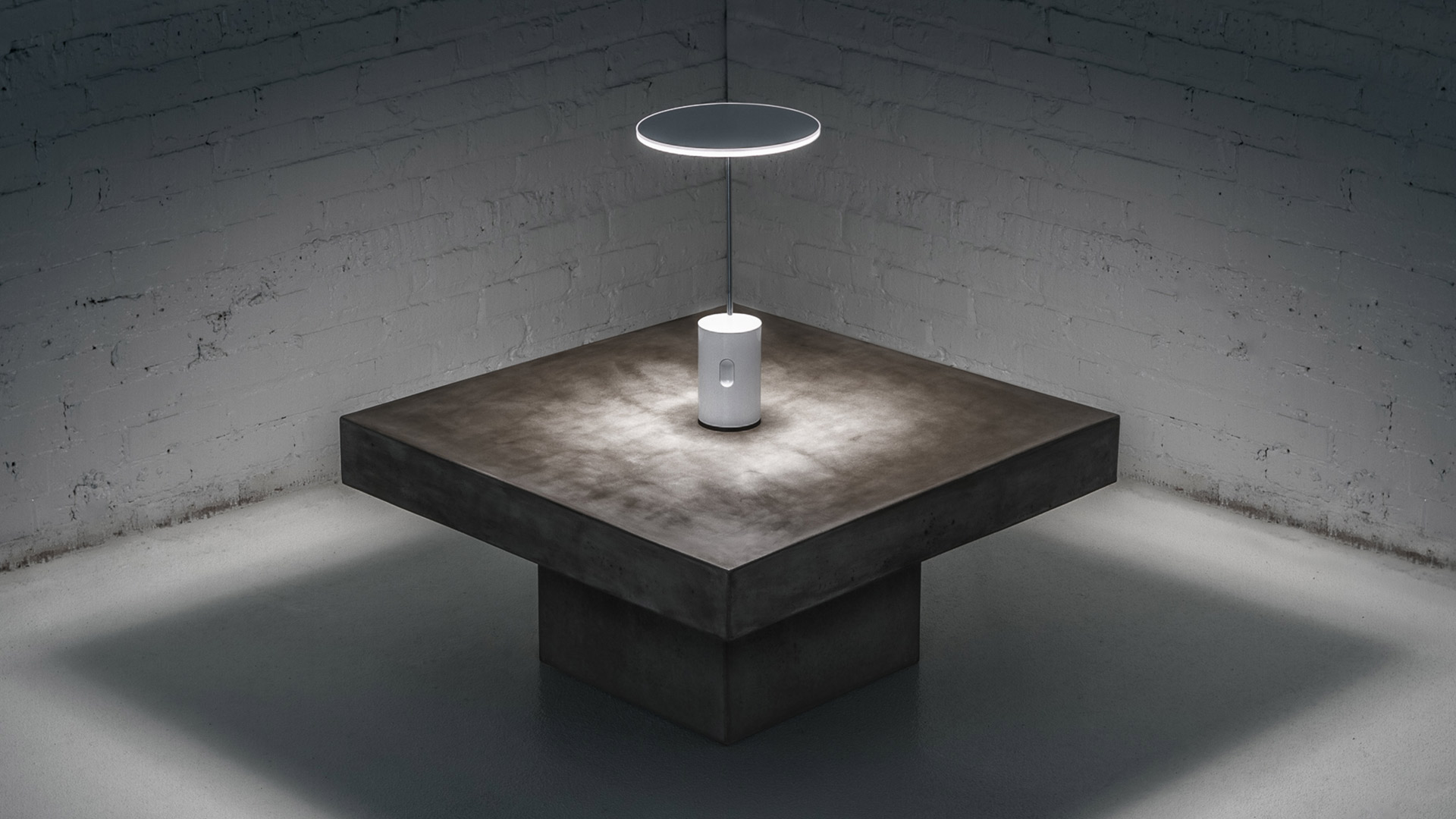 Artemide Sisifo portable lamp on concrete table beneath a circular pendant light in a minimalist grey brick room