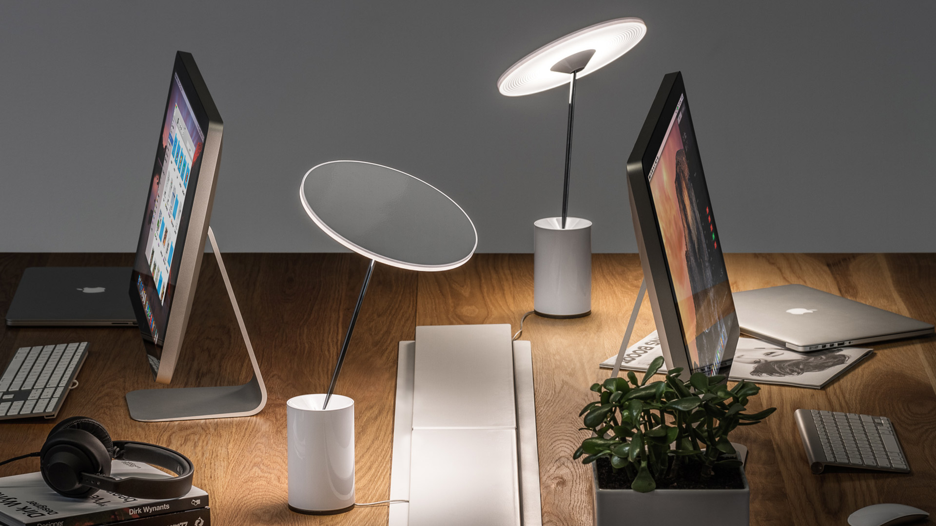 Two Artemide Sisifo table lamps with circular disc shades on cylindrical white bases, placed on a desk with monitors and accessories