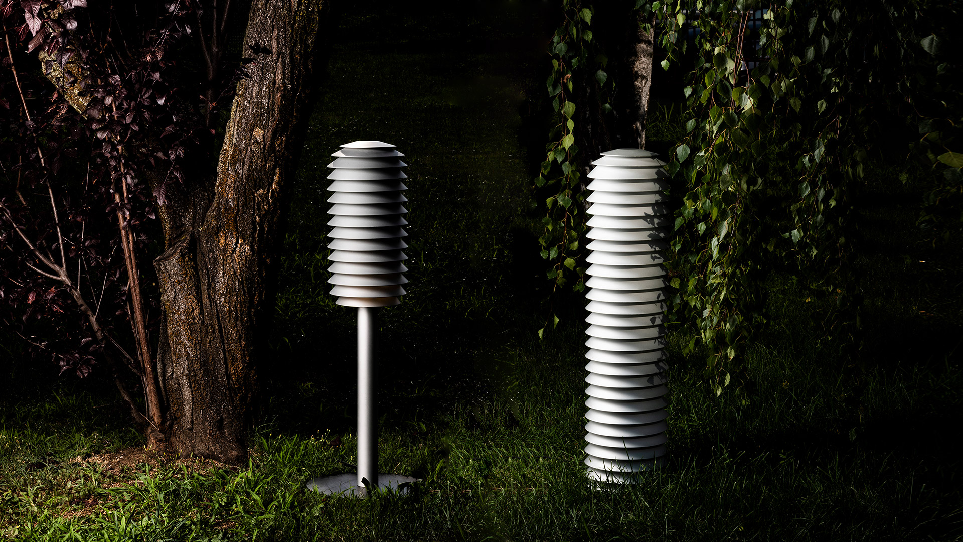 Due lampade da esterno Artemide Slicing con paralumi a dischi bianchi sovrapposti: una su palo, una a terra, che illuminano un giardino di notte