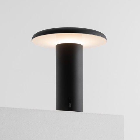 Artemide Takku portable table lamp in black, featuring a cylindrical stem and flat circular diffuser emitting warm white light