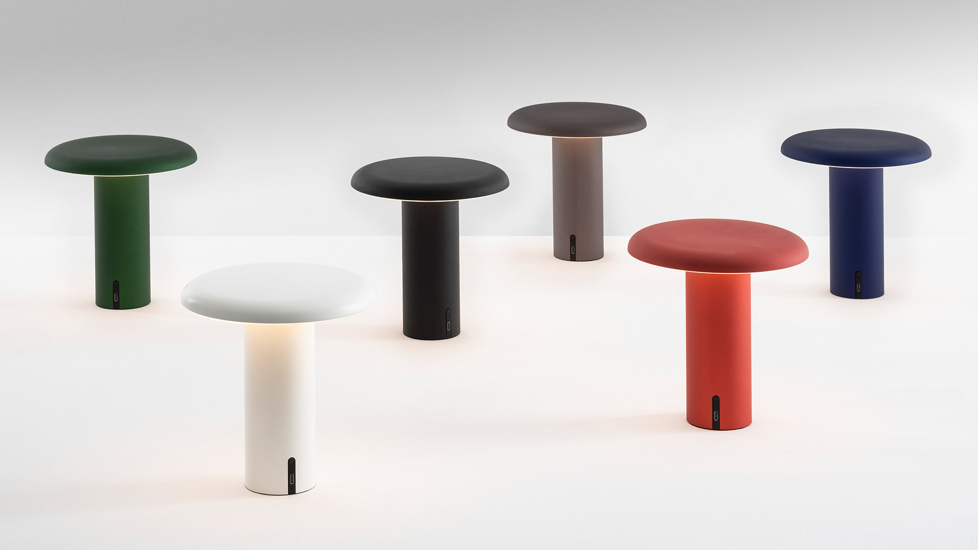 Artemide Takku portable LED table lamp with white mushroom-shaped diffuser, dark cylindrical body, and circular base, shown next to an apple for scale