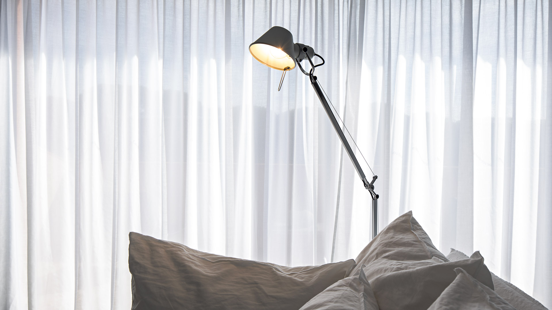 Lampada da terra Artemide Tolomeo Terra con paralume scuro e braccio articolato, posizionata accanto a cuscini davanti a tende bianche