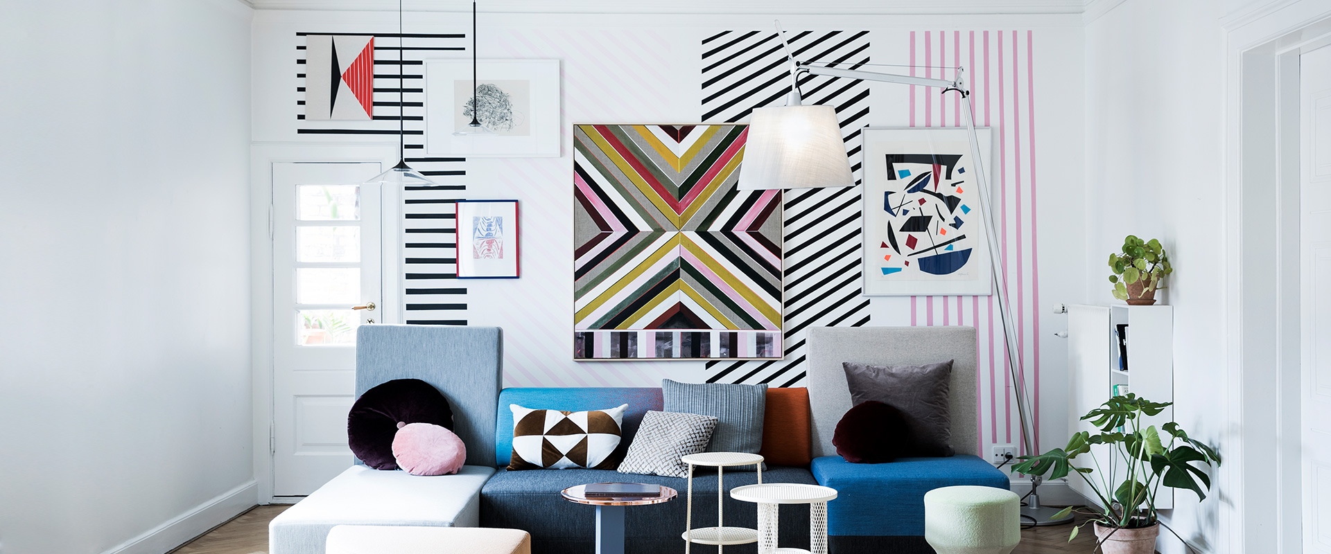 Soggiorno con arte murale geometrica vivace, divano componibile colorato e lampada da terra Artemide Tolomeo Maxi con paralume bianco
