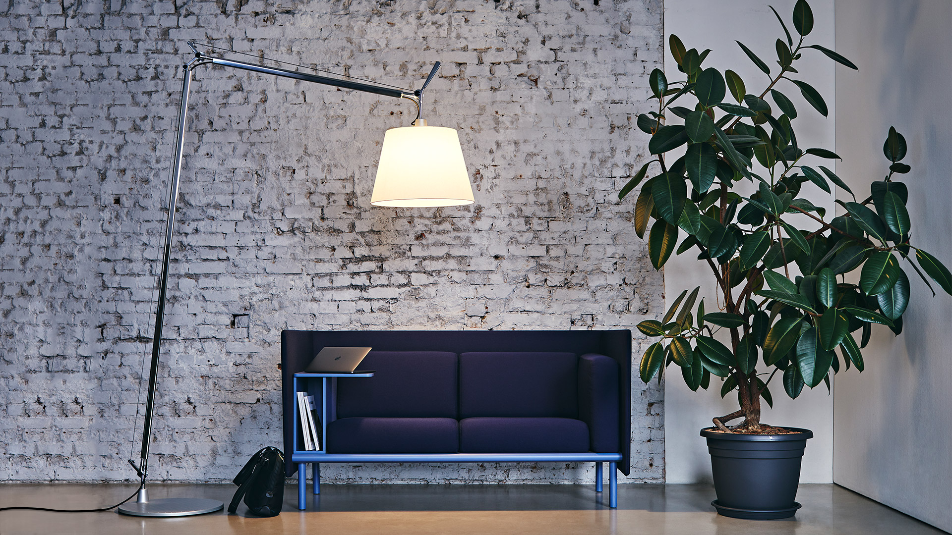 Lampada da terra Artemide Tolomeo Maxi con paralume bianco che illumina un divano scuro davanti a un muro di mattoni bianchi con pianta in vaso