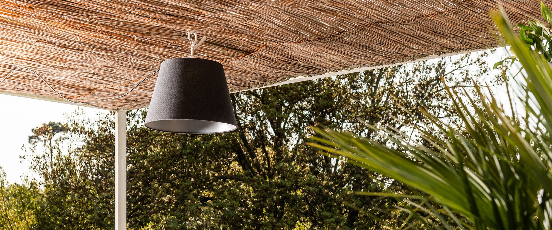 Lampada a sospensione Tolomeo Paralume Outdoor con paralume conico nero appesa a una pergola in cannuccia naturale, circondata da rigogliosa vegetazione