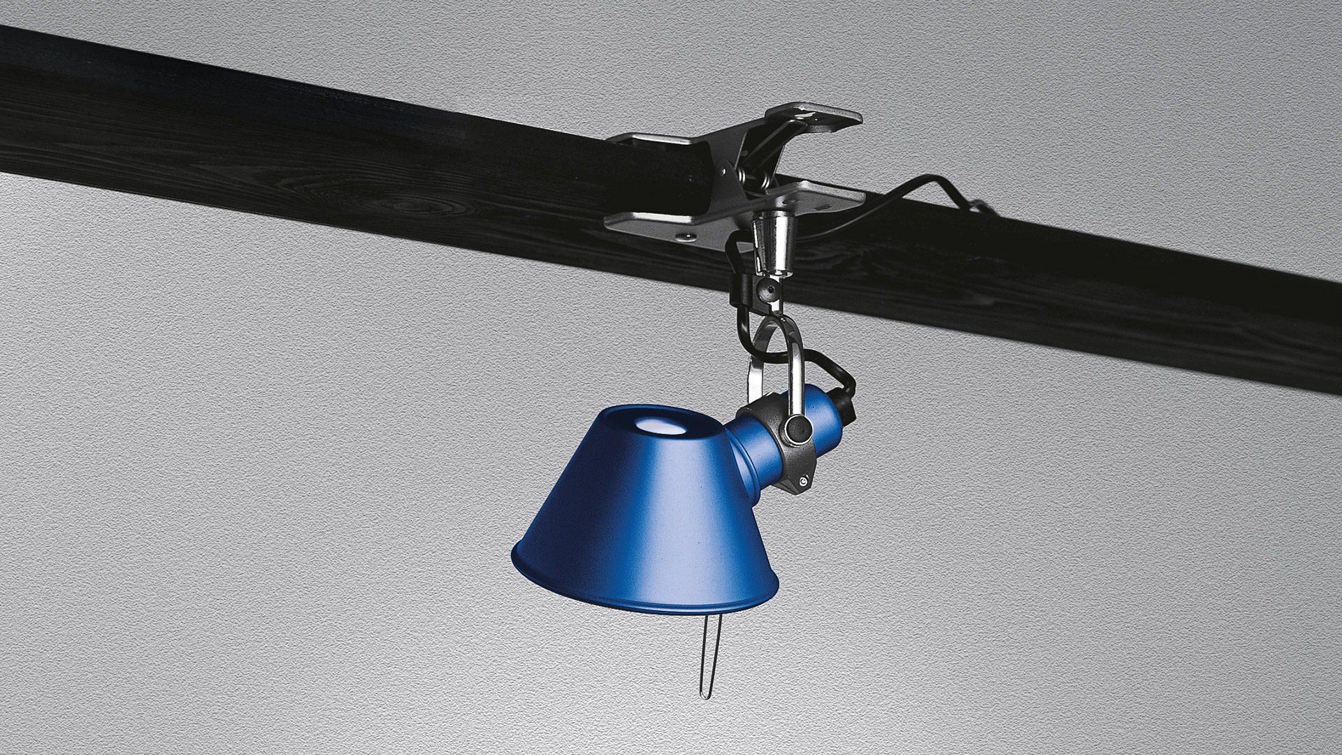 Lampada Artemide Tolomeo Pinza in blu, agganciata a una trave scura, con paralume conico rivolto verso il basso