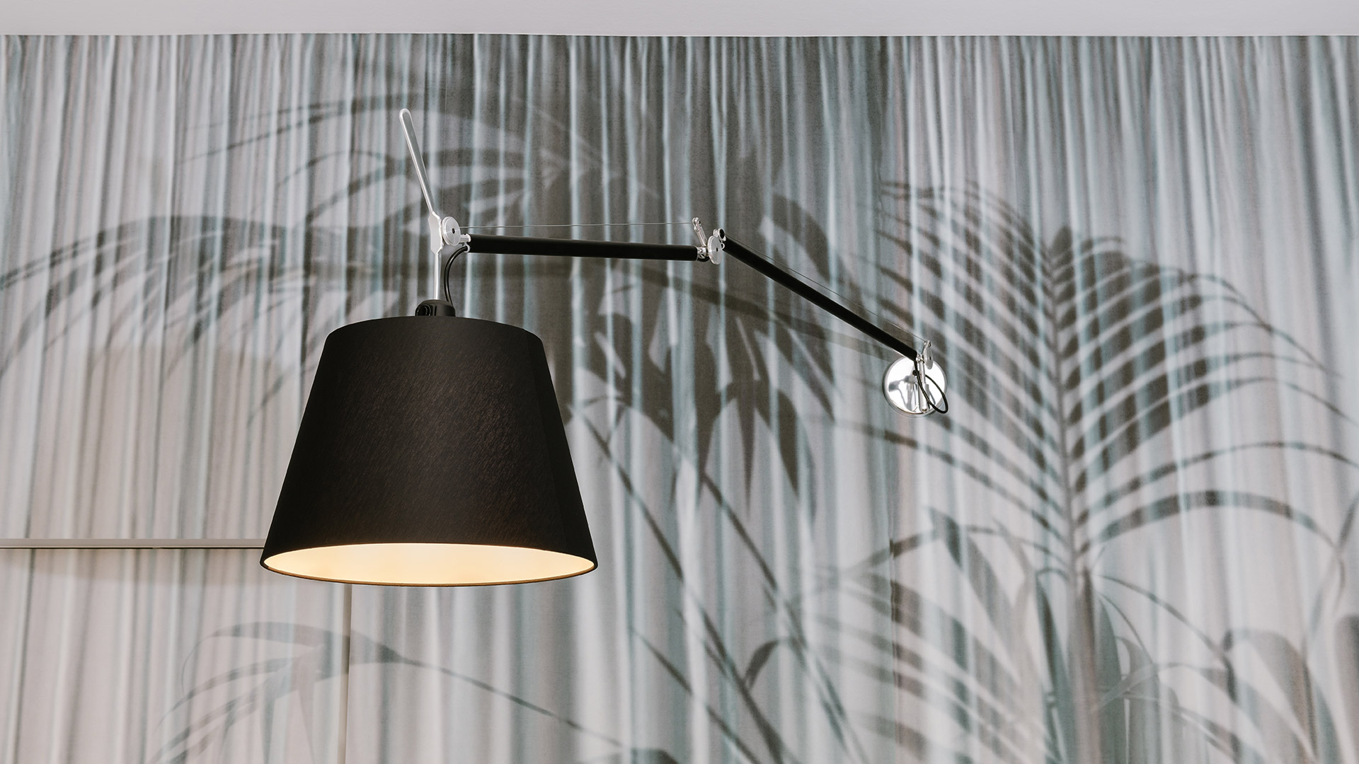 Lampada da parete Artemide Tolomeo Mega Parete con paralume in tessuto nero e braccio in alluminio regolabile, montata su tenda con motivo a foglie tropicali