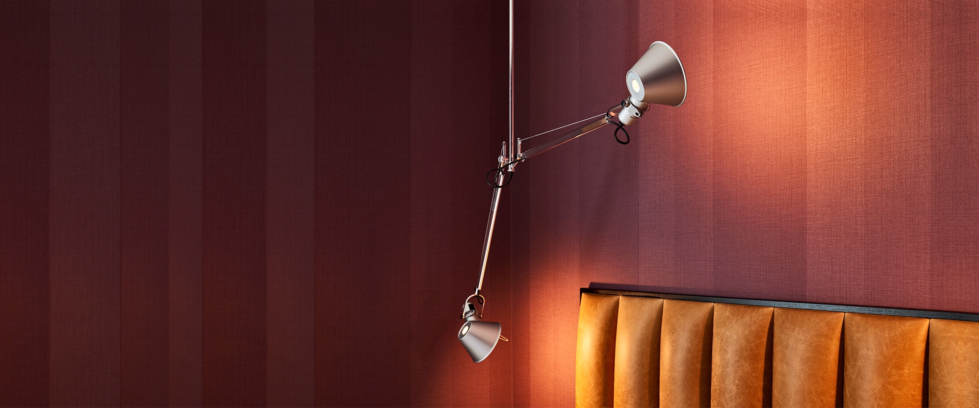 Tolomeo Due Bracci Sospensione: due lampade a sospensione regolabili in alluminio con bracci articolati, montate a parete sopra divano in pelle arancione