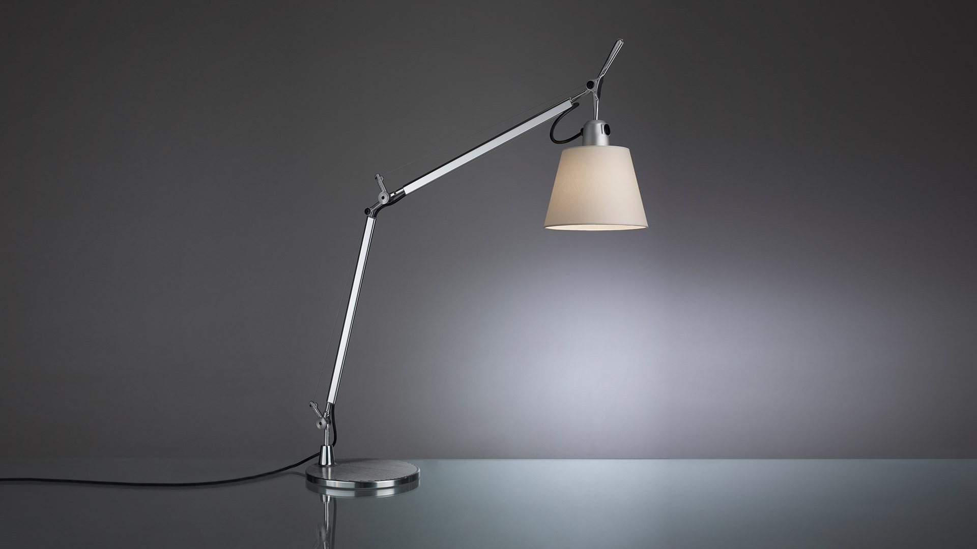 Artemide Tolomeo Basculante Tavolo desk lamp with adjustable aluminum arm, conical fabric shade, and circular base on reflective surface