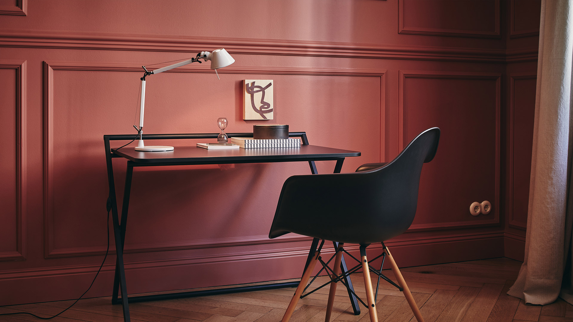 Artemide Tolomeo desk lamp on a minimalist black desk with a black chair, set against a warm red paneled wall.