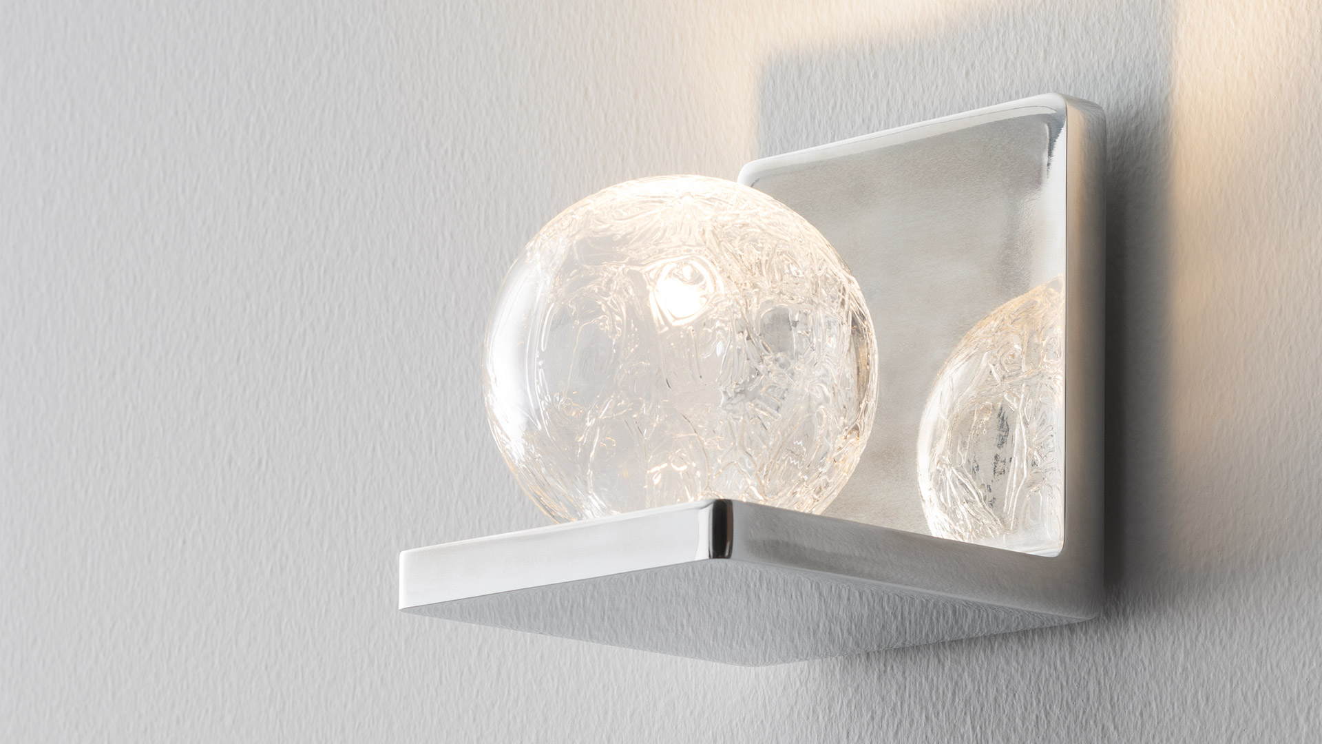Due applique da parete Artemide Ulrich und Diotima: sinistra in alluminio argento, destra in finitura marrone, entrambe emettono luce calda verso l'alto.