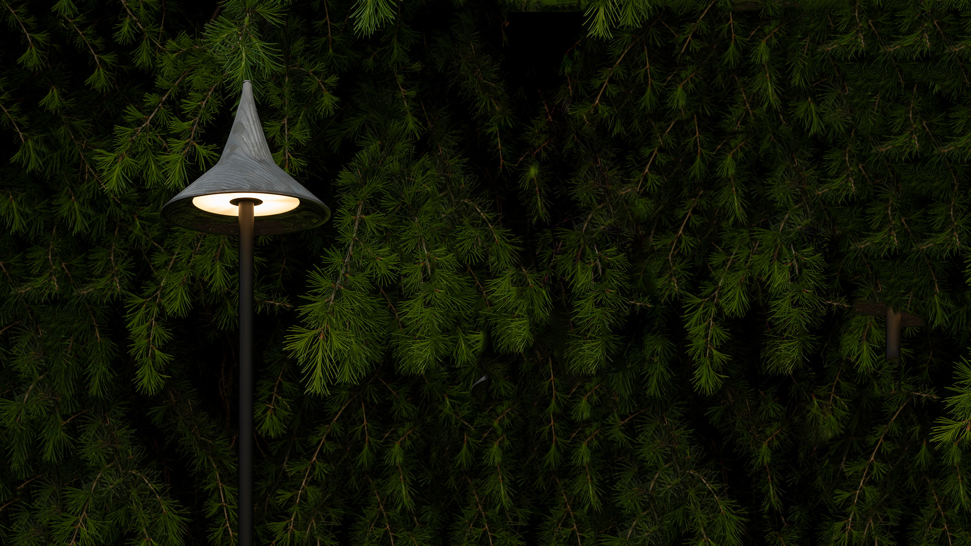 Lampada da palo Artemide Unterlinden Outdoor con paralume conico che emette luce calda, su sfondo di siepe sempreverde al crepuscolo