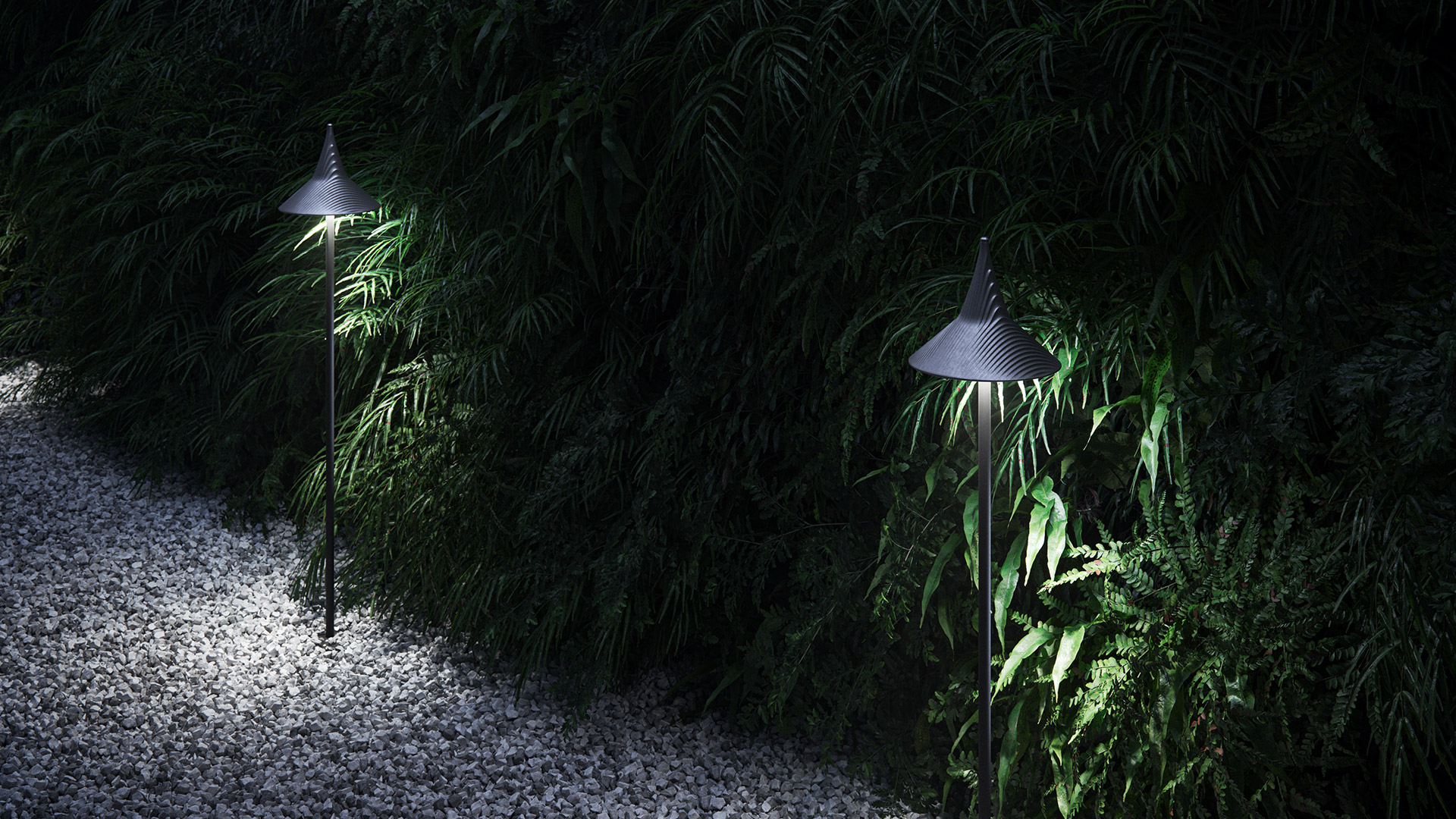 Due lampade da esterno Artemide Unterlinden con paralumi conici scuri che illuminano un vialetto di ghiaia bordato da rigogliosa vegetazione di notte