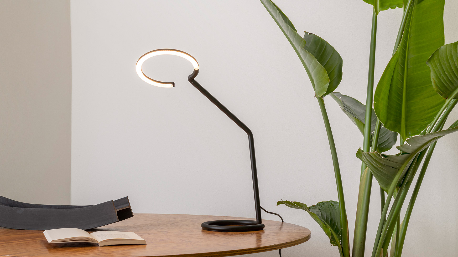 Artemide Vine Light desk lamp in black with circular LED head on a curved arm, placed on a white desk beside a laptop and notepad